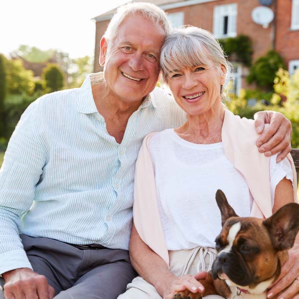Senioren-Paar mit Hund, das im Garten sitzt und zusammen in die Kamera blickt