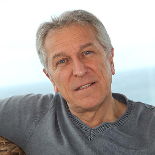 älterer Herr mit grauen Haaren, blauen Augen und grauem Pullover, der selbstbewusst und direkt in die Kamera blickt