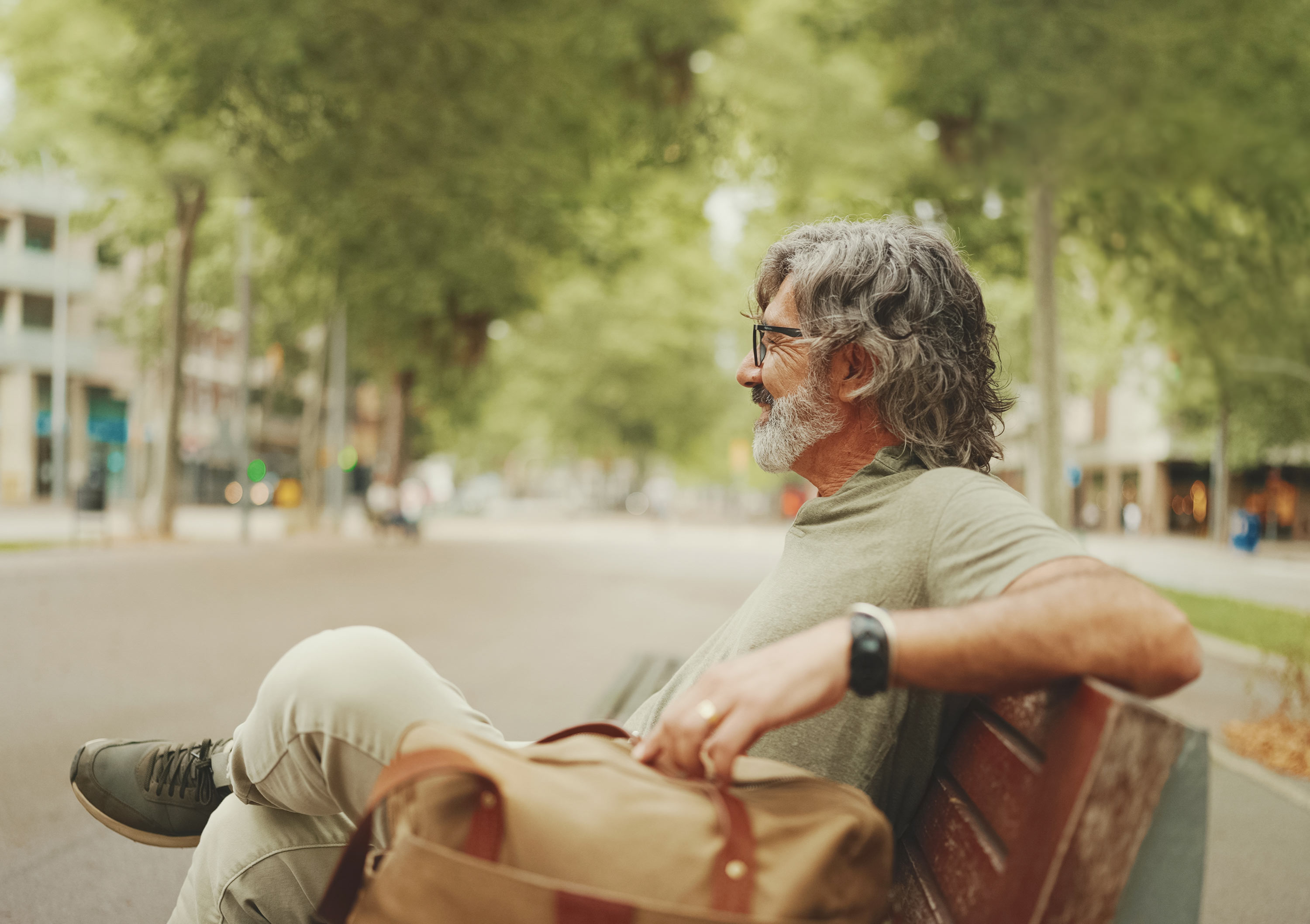Positive middle-aged man with gray hair and beard wearing casual clothes sits on bench. Mature gentleman in eyeglasses is resting on bench