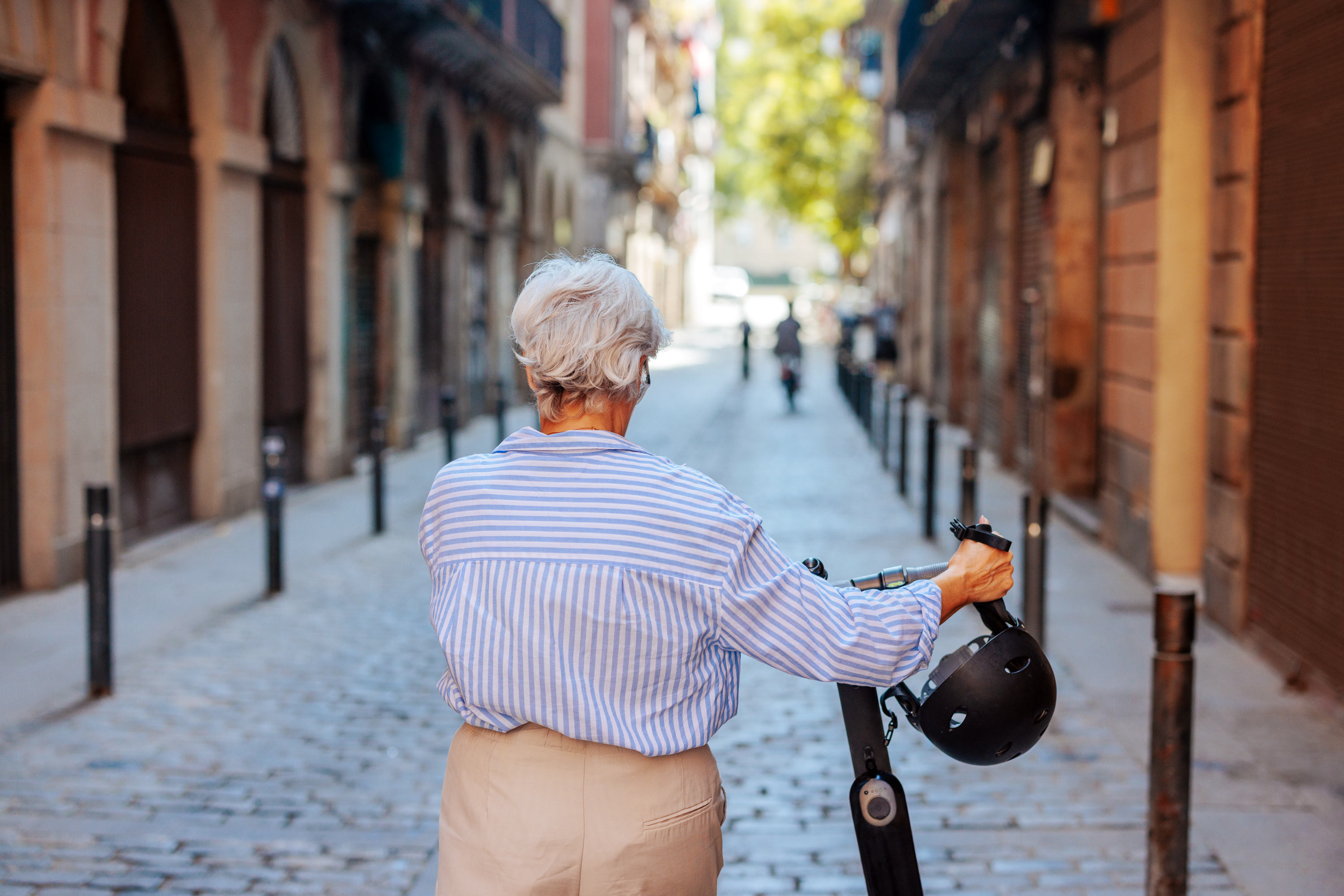 A senior Caucasian woman is pictured from the rear, walking down the street with her electric scooter next to her.